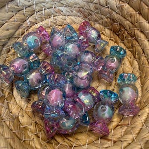 Puede incluir: Una colección de cuentas iridiscentes en forma de caramelo, en tonos rosa, azul y morado, dentro de una cesta tejida. Las cuentas tienen un acabado brillante y están dispuestas al azar, creando una exhibición colorida y juguetona.