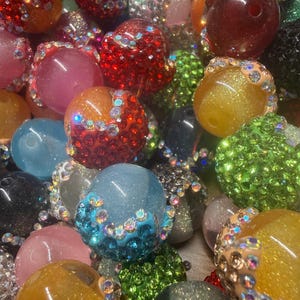 May include: A collection of colorful, sparkling beads in various sizes and hues. The beads are adorned with rhinestones and glitter, creating a dazzling effect. Colors include red, pink, blue, green, and gold. These beads are suitable for jewelry making and crafts.