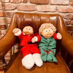 May include: Two Cabbage Patch Kids dolls dressed in holiday outfits. One doll wears a red Santa suit with white trim, and the other wears a green elf outfit. Both dolls have curly hair and are seated on a brown leather chair.