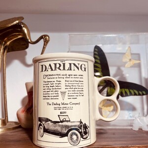 May include: Cream-colored ceramic mug featuring a vintage car advertisement for The Darling Motor Company from 1917. The mug has a unique handle with two finger holes and includes text and a car illustration.