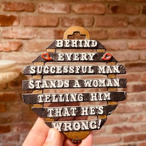 May include: A decorative sign with the text "BEHIND EVERY SUCCESSFUL MAN STANDS A WOMAN TELLING HIM THAT HE'S WRONG!" The sign is black and gold with white lettering and red accents. The background is a brick wall.