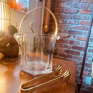 May include: A clear glass ice bucket with a gold-colored handle and a pair of matching ice tongs. The ice bucket has a square base and a cylindrical body. The tongs have decorative fan-shaped ends.