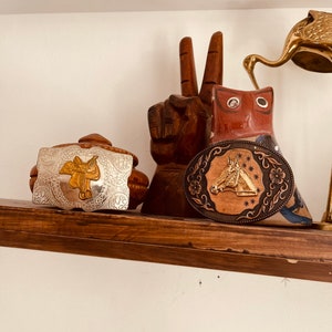 May include: A shelf displays decorative items, including a silver and gold belt buckle with a saddle design, a wooden peace sign, a carved owl, and a bronze horse head belt buckle. The items are arranged on a wooden shelf.