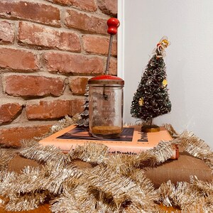 May include: A vintage red-topped food chopper with a glass jar, sitting on a stack of books. A small Christmas tree with a gold angel topper and gold tinsel garland are also present. The background features a brick wall and a white wall.