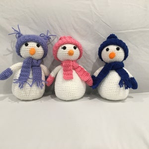 Op de afbeelding: Drie gehaakte sneeuwpoppen met witte lichamen, oranje wortelneuzen en zwarte knoopogen. Elke sneeuwpop draagt een andere kleur hoed en sjaal: paars, roze en blauw. Handgemaakt en geschikt voor winterdecoratie.
