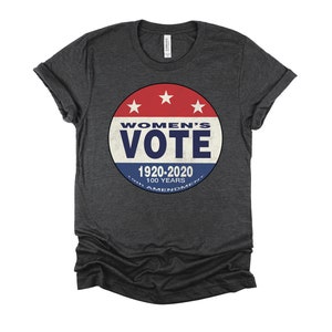 May include: A dark gray t-shirt with a red, white, and blue circular graphic that says "Women's Vote" with the text "1920-2020 100 Years 19th Amendment" underneath.