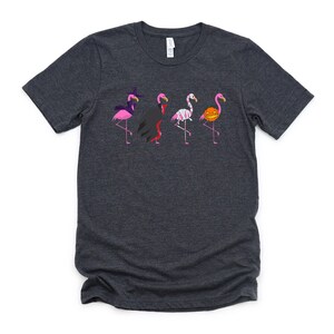 May include: A dark gray t-shirt with a graphic of five pink flamingos dressed in Halloween costumes. The flamingos are wearing a witch hat, a vampire cape, a mummy costume, a pumpkin on their head, and a witch hat.