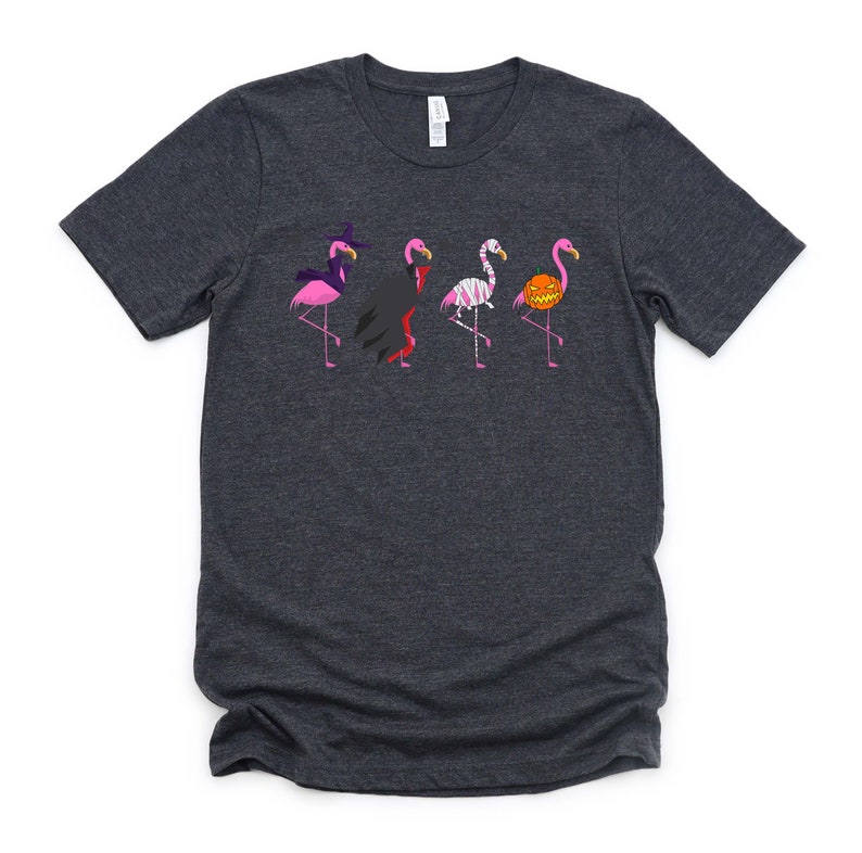 May include: A dark gray t-shirt with a graphic of five pink flamingos dressed in Halloween costumes. The flamingos are wearing a witch hat, a vampire cape, a mummy costume, a pumpkin on their head, and a witch hat.