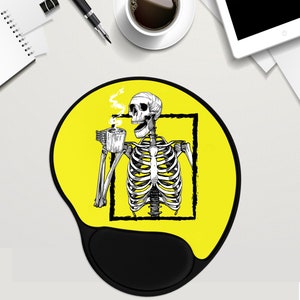 May include: A yellow mouse mat with a black skeleton drinking coffee. The skeleton is inside a black square frame. The mouse mat has a black wrist rest.
