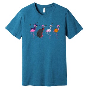 May include: A teal blue t-shirt with a graphic of five pink flamingos dressed in Halloween costumes. The flamingos are wearing a witch hat, a vampire cape, a mummy costume, a pumpkin, and a witch hat.