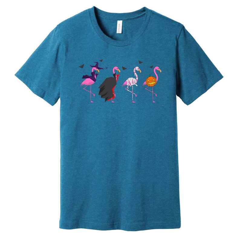 May include: A teal blue t-shirt with a graphic of five pink flamingos dressed in Halloween costumes. The flamingos are wearing a witch hat, a vampire cape, a mummy costume, a pumpkin, and a witch hat.