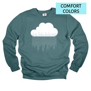 Op de afbeelding: Tealkleurige crewneck sweatshirt met een witte wolkengrafiek met binaire code als regen. De sweatshirt heeft lange mouwen en een relaxte pasvorm. De tekst "COMFORT COLORS" staat in een lichtblauwe rechthoek.