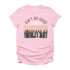 Pu&ograve; includere: Una maglietta rosa chiaro con maniche arrotolate. La maglietta presenta il testo "Ain't No Hood Like Sainthood" in oro e bianco, con un'illustrazione di santi sottostante. La maglietta &egrave; realizzata in un tessuto morbido.