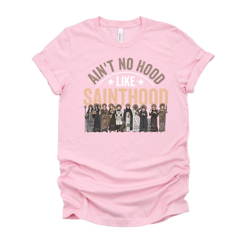 Pu&ograve; includere: Una maglietta rosa chiaro con maniche arrotolate. La maglietta presenta il testo "Ain't No Hood Like Sainthood" in oro e bianco, con un'illustrazione di santi sottostante. La maglietta &egrave; realizzata in un tessuto morbido.