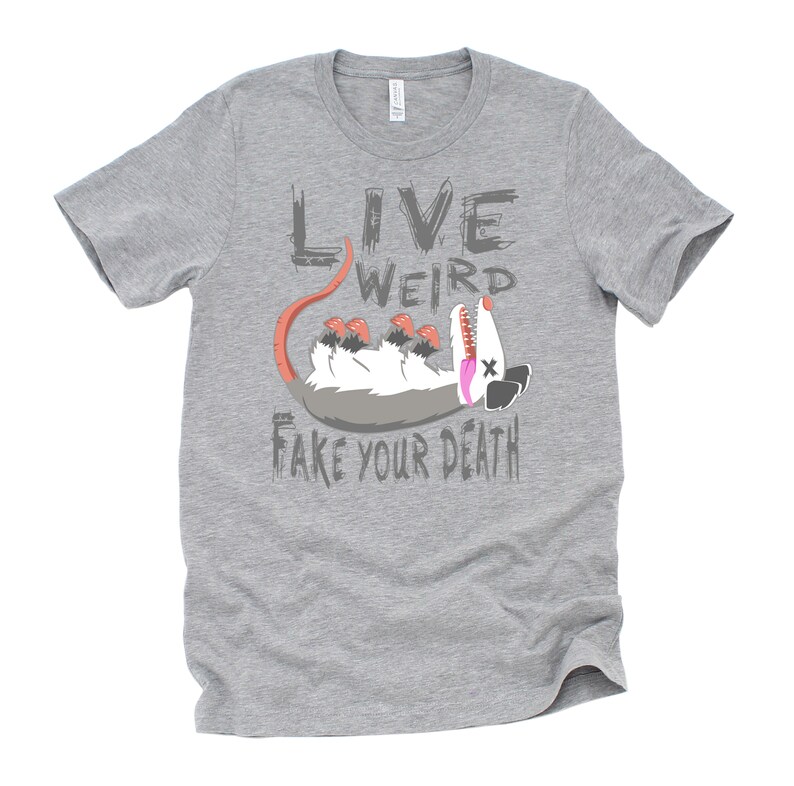 Op de afbeelding: Grijs T-shirt met een cartoon opossum en de tekst "Live Weird Fake Your Death".