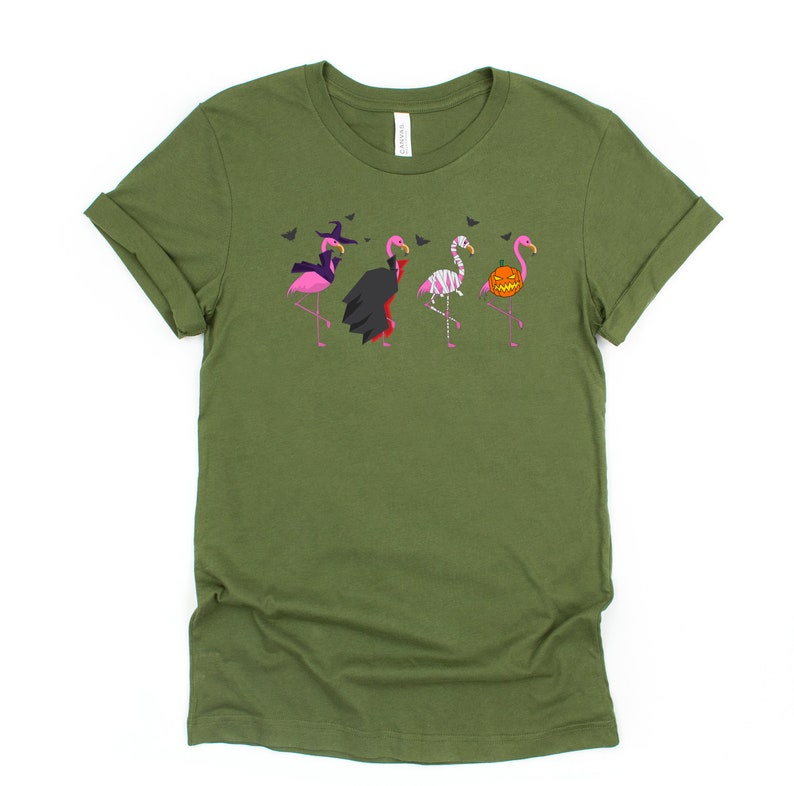 May include: A green t-shirt with a graphic of five pink flamingos dressed in Halloween costumes. The flamingos are wearing a witch hat, a vampire cape, a mummy costume, a skeleton costume, and a pumpkin head.