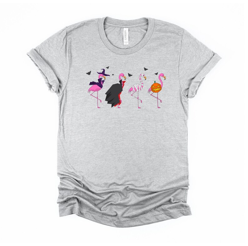 May include: A gray t-shirt with a graphic of four pink flamingos dressed in Halloween costumes. The flamingos are dressed as a witch, a vampire, a mummy, and a pumpkin.