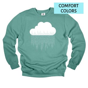 Op de afbeelding: Tealkleurige sweatshirt met een witte wolkengrafiek en binaire code regen. De tekst "COMFORT COLORS" staat in een lichtblauwe rechthoek. De sweatshirt heeft lange mouwen en een ronde hals.