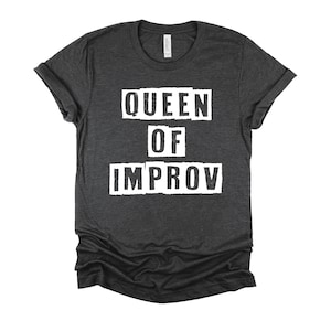 May include: Dark heather grey t-shirt with white text that reads "QUEEN OF IMPROV" in a distressed font.