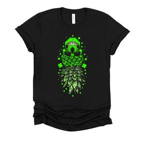 May include: A black short-sleeve t-shirt featuring a central graphic of a bright green skull resting atop a pineapple, surrounded by scattered four-leaf clover icons. The design is rendered in vibrant neon green ink against the dark fabric.