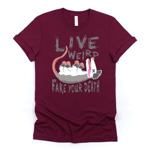 Op de afbeelding: Een maroon t-shirt met een grijze afbeelding van een opossum die dood speelt en de tekst "Live Weird Fake Your Death".