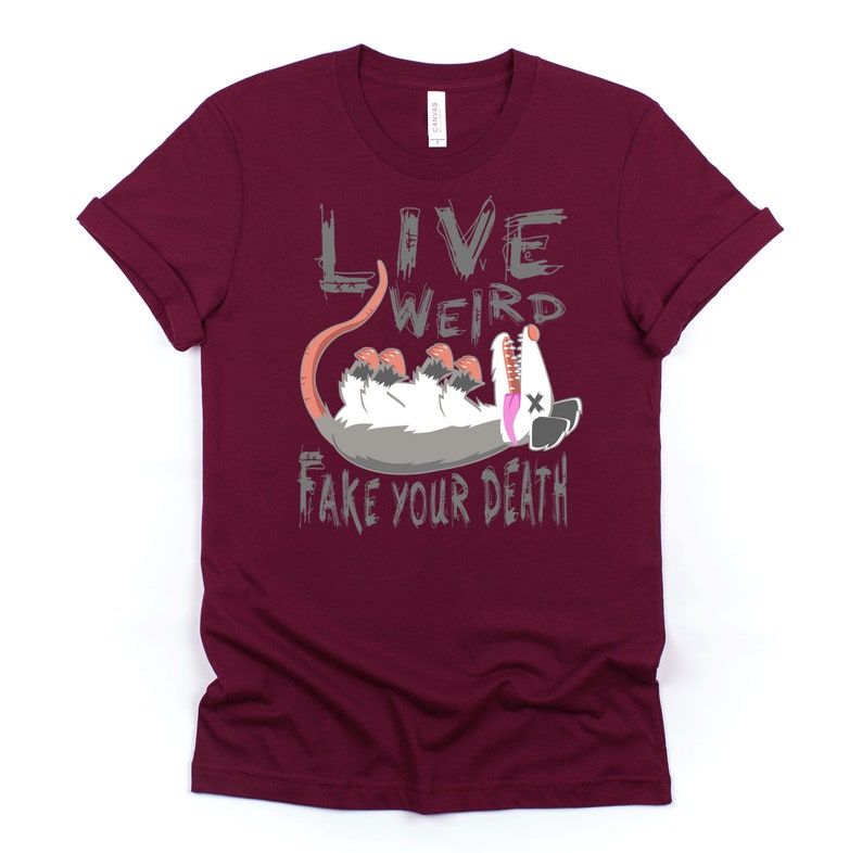 Op de afbeelding: Een maroon t-shirt met een grijze afbeelding van een opossum die dood speelt en de tekst "Live Weird Fake Your Death".