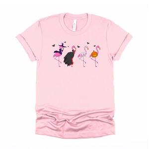 May include: Pink t-shirt with a graphic of four flamingos dressed in Halloween costumes. The flamingos are dressed as a witch, a vampire, a mummy, and a pumpkin.