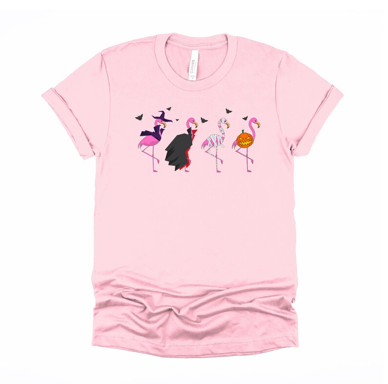 May include: Pink t-shirt with a graphic of four flamingos dressed in Halloween costumes. The flamingos are dressed as a witch, a vampire, a mummy, and a pumpkin.