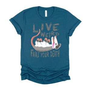 Op de afbeelding: Tealblauwe T-shirt met een grijze afbeelding van een opossum met de tekst "Live Weird Fake Your Death".