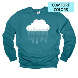 Op de afbeelding: Teal sweatshirt met een witte wolkengrafiek en binaire code regen. De tekst "COMFORT COLORS" staat in een lichtblauwe rechthoek. De sweatshirt heeft lange mouwen.