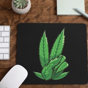 May include: Black mousepad featuring a stylized green cannabis leaf design. The mousepad is rectangular and sits on a wooden surface, alongside a keyboard, mouse, pen, and notepad.