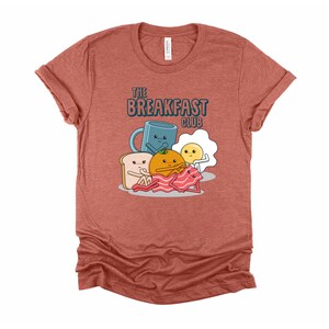 May include: A heather red t-shirt featuring a cartoon breakfast club graphic. The graphic depicts a mug, an egg, a slice of toast, an orange, and a piece of bacon. The text "The Breakfast Club" is printed above the graphic.