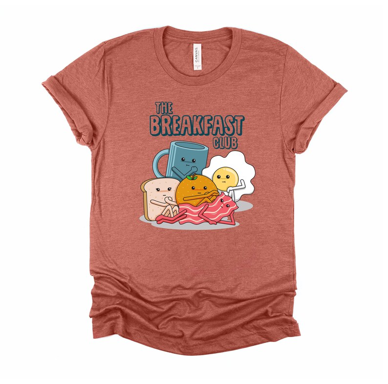 May include: A heather red t-shirt featuring a cartoon breakfast club graphic. The graphic depicts a mug, an egg, a slice of toast, an orange, and a piece of bacon. The text "The Breakfast Club" is printed above the graphic.