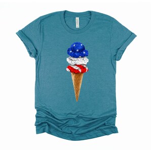 May include: A teal blue t-shirt with a graphic of a three-scoop ice cream cone. The ice cream is decorated with the colors of the American flag: red, white, and blue.