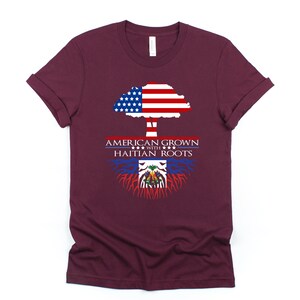 May include: A maroon t-shirt with a tree design. The tree is made up of the American flag and has roots made up of the Haitian flag. The text "AMERICAN GROWN WITH HAITIAN ROOTS" is printed on the shirt.