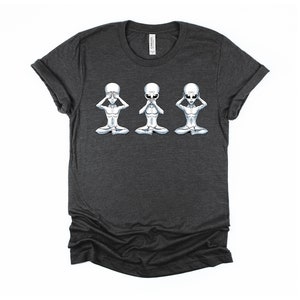 May include: Dark heather grey t-shirt featuring a graphic of three grey aliens in a row, each with one hand covering their eyes, one hand covering their mouth, and one hand covering their ears. The aliens are in a seated position with their legs crossed.