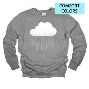 Op de afbeelding: Een grijze sweatshirt met een witte wolkengrafiek en binaire code regen. De tekst "COMFORT COLORS" staat in een lichtblauwe rechthoek. De sweatshirt heeft lange mouwen en een ronde hals.