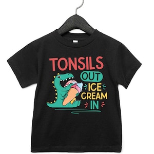 May include: Black t-shirt with a green dinosaur eating ice cream. The text on the shirt reads "Tonsils Out Ice Cream In".