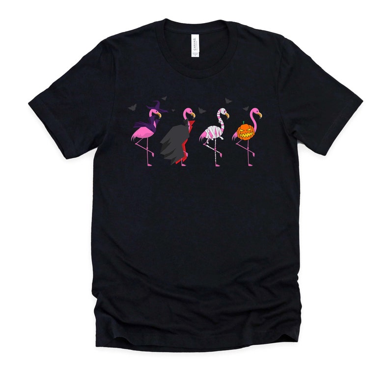 May include: A black t-shirt with a graphic of five pink flamingos dressed in Halloween costumes. The flamingos are wearing a witch hat, a vampire cape, a mummy costume, a skeleton costume, and a pumpkin costume.
