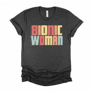 May include: A dark heather grey t-shirt with the text "Bionic Woman" in colorful retro-style lettering.