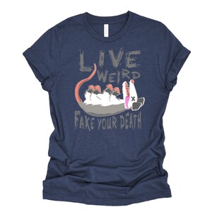 Op de afbeelding: Een marineblauw t-shirt met een grijze afbeelding van een opossum die dood speelt. De tekst "Live Weird Fake Your Death" is in grijs op het t-shirt gedrukt.