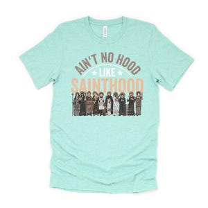 Pu&ograve; includere: Una maglietta verde acqua con la scritta "AIN'T NO HOOD LIKE SAINTHOOD" in marrone e pesca. Sotto il testo c'&egrave; una grafica di diverse figure in abiti religiosi. La maglietta &egrave; realizzata in un materiale morbido.