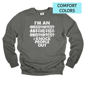 May include: A dark gray sweatshirt with the text "I'M AN ANEESTHATIST ANYTHETIST ANISTHATIST I KNOCK PEOPLE OUT" in white. The words "ANEESTHATIST", "ANYTHETIST", and "ANISTHATIST" are crossed out. The top right corner has a light blue rectangle with the words "COMFORT COLORS".