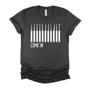 May include: A dark heather grey t-shirt with a white graphic of lock picking tools and the text "COME IN".