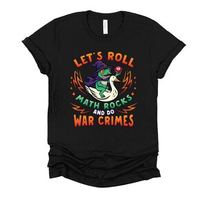 May include: Black t-shirt with a colorful graphic featuring a frog wearing a wizard hat and cape, riding a swan. Text reads "LET'S ROLL MATH ROCKS AND DO WAR CRIMES". The design includes a red die and stars.
