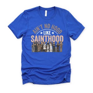 Pu&ograve; includere: T-shirt blu reale con la scritta "Ain't No Hood Like Sainthood" in un design circolare. Il design include un'illustrazione di figure in abiti religiosi, aggiungendo un tocco unico all'abbigliamento.