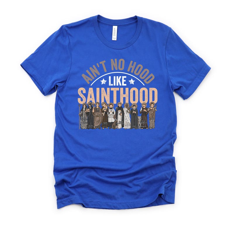 Pu&ograve; includere: T-shirt blu reale con la scritta "Ain't No Hood Like Sainthood" in un design circolare. Il design include un'illustrazione di figure in abiti religiosi, aggiungendo un tocco unico all'abbigliamento.