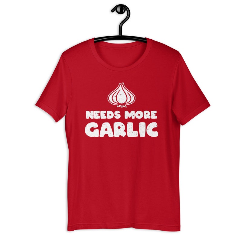 May include: Red short-sleeved t-shirt with a white graphic of a garlic clove and the text "Needs More Garlic".