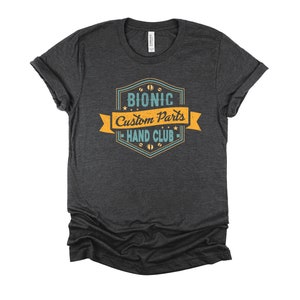 May include: A dark heather grey t-shirt with a vintage-style logo that reads "Bionic Custom Parts Hand Club". The logo is in shades of blue and yellow with a yellow banner.