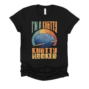 May include: Black t-shirt with a vintage-style graphic of a ball of yarn and knitting needles. The text "I'm a Knotty Knotty Hooker" is printed on the shirt.